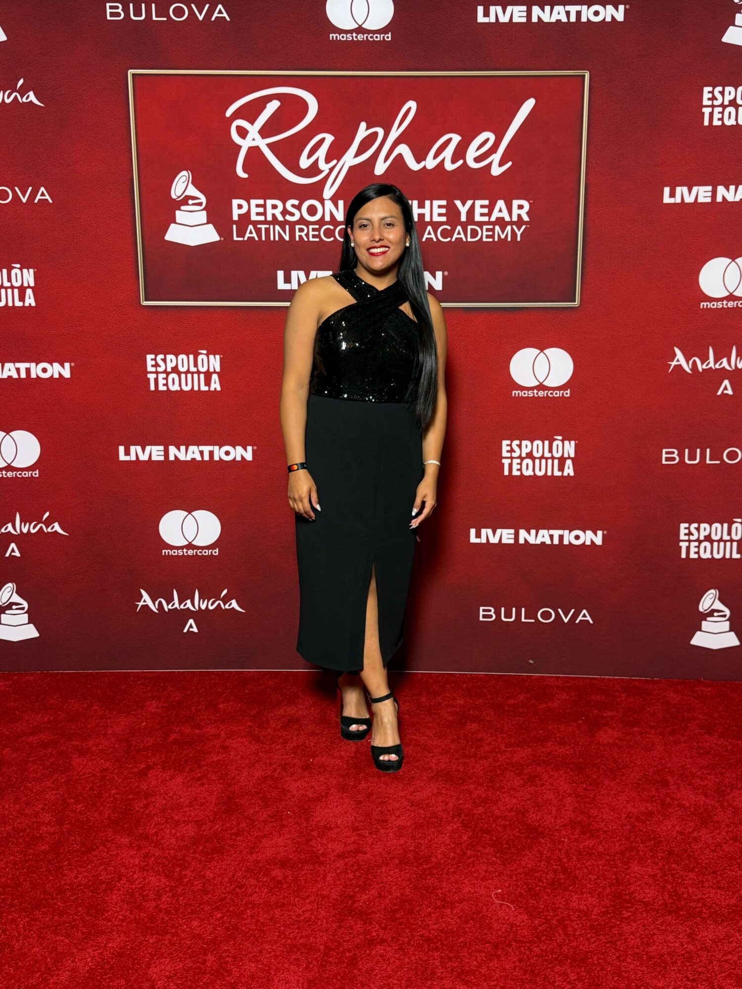 Woman in black dress standing on red carpet with event backdrop behind her, smiling, with her hands at her sides.