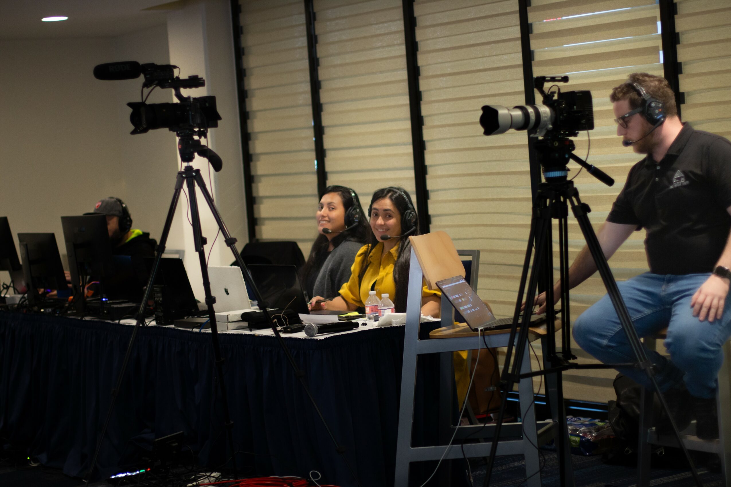 Three women sit at a table with cameras and equipment, a man stands nearby in a room with large window blinds.