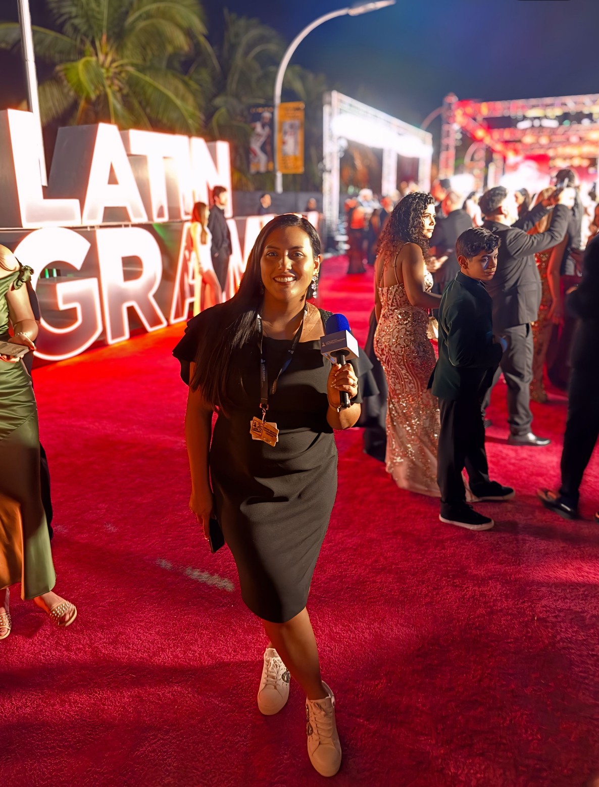 Woman standing on red carpet at event, holding microphone, smiling, with large illuminated sign in background, night scene.