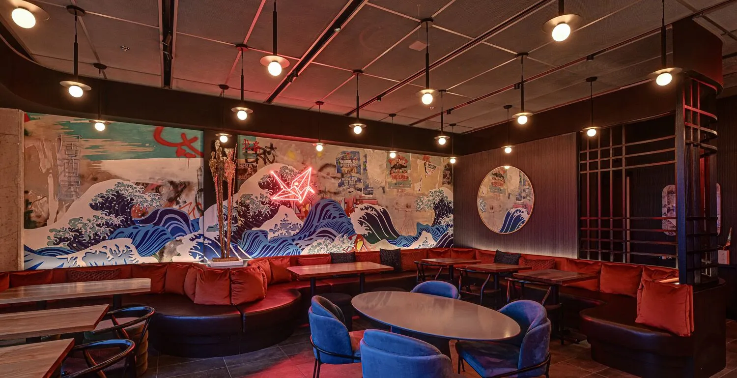Interior of a restaurant with a mural, round tables, and colorful chairs, illuminated by ceiling lights.