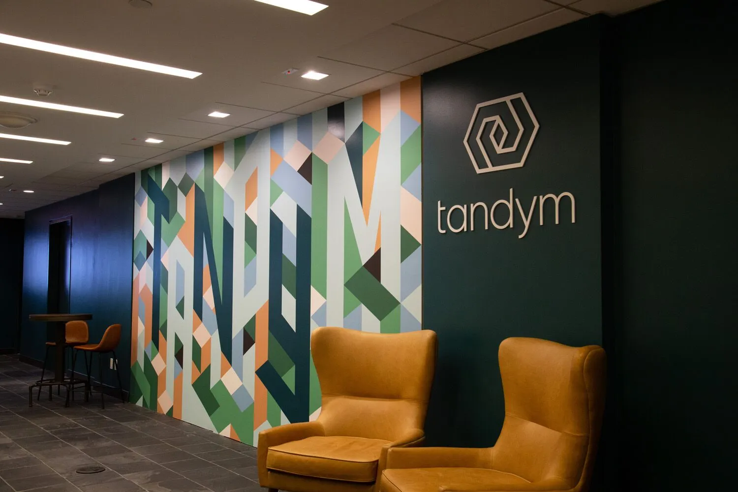 Interior office wall with colorful geometric pattern, logo, and the word 'tandym', with two yellow chairs in front.