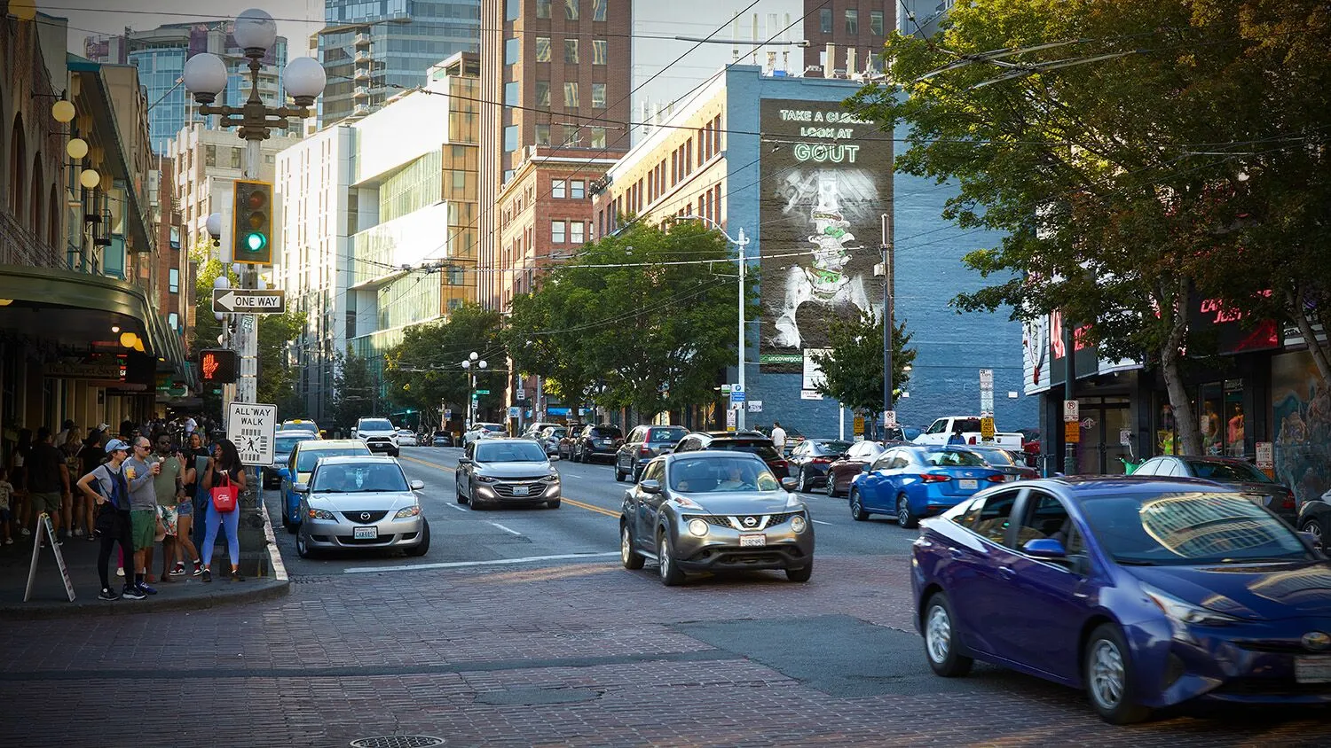 City street with cars, pedestrians, tall buildings, and trees, during daytime with clear weather.