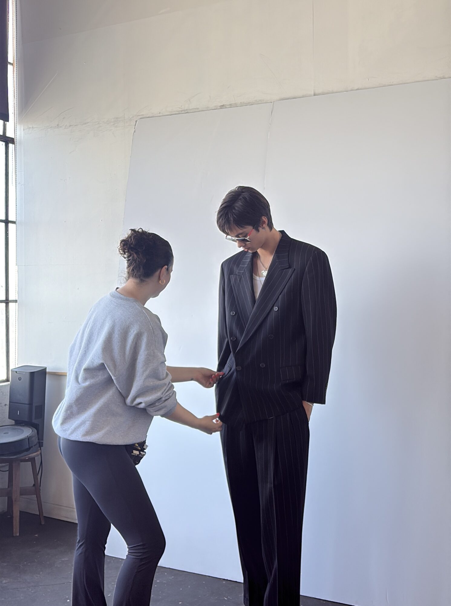 Two people standing and interacting indoors, one in a white top and the other in a dark suit, against a plain wall.
