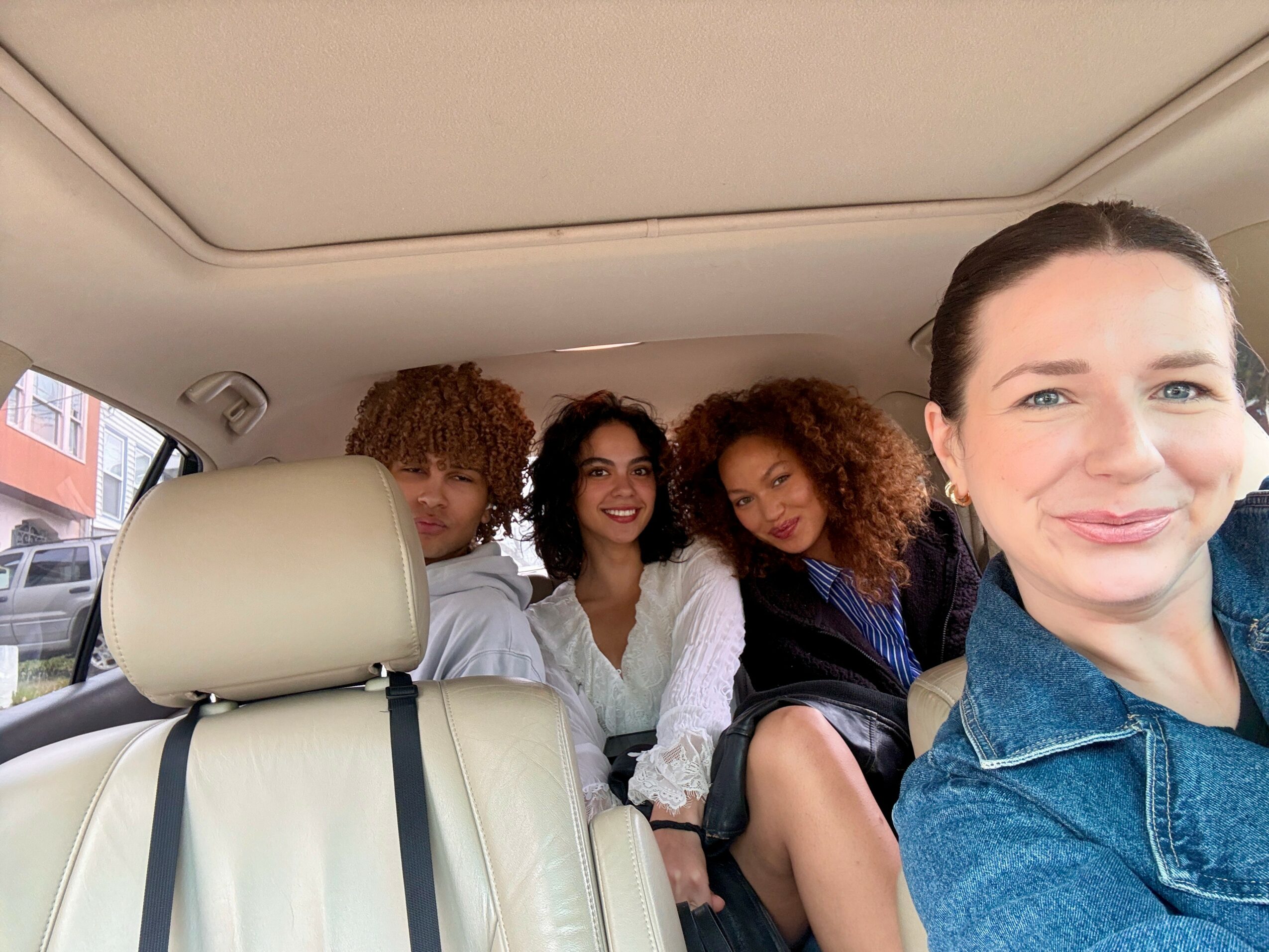 Four women sitting in a car, smiling, with the woman on the right taking a selfie. The car interior is visible.