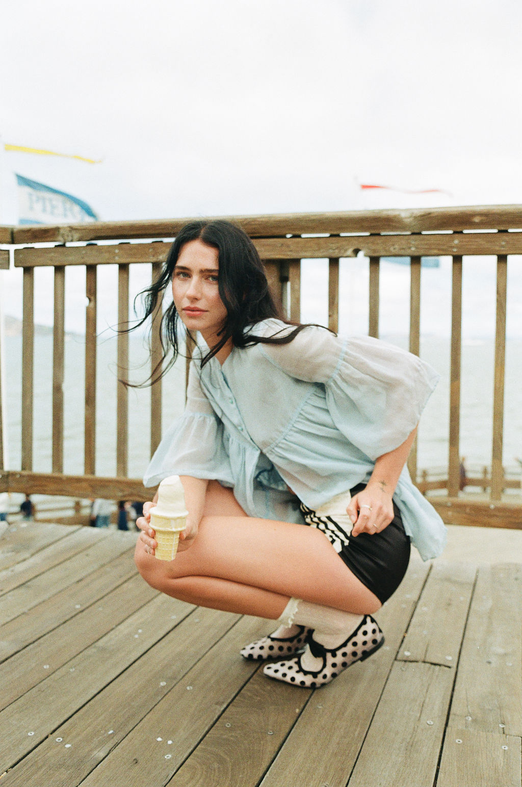 Young woman crouching on a wooden deck holding an ice cream cone, with a railing and cloudy sky in background.