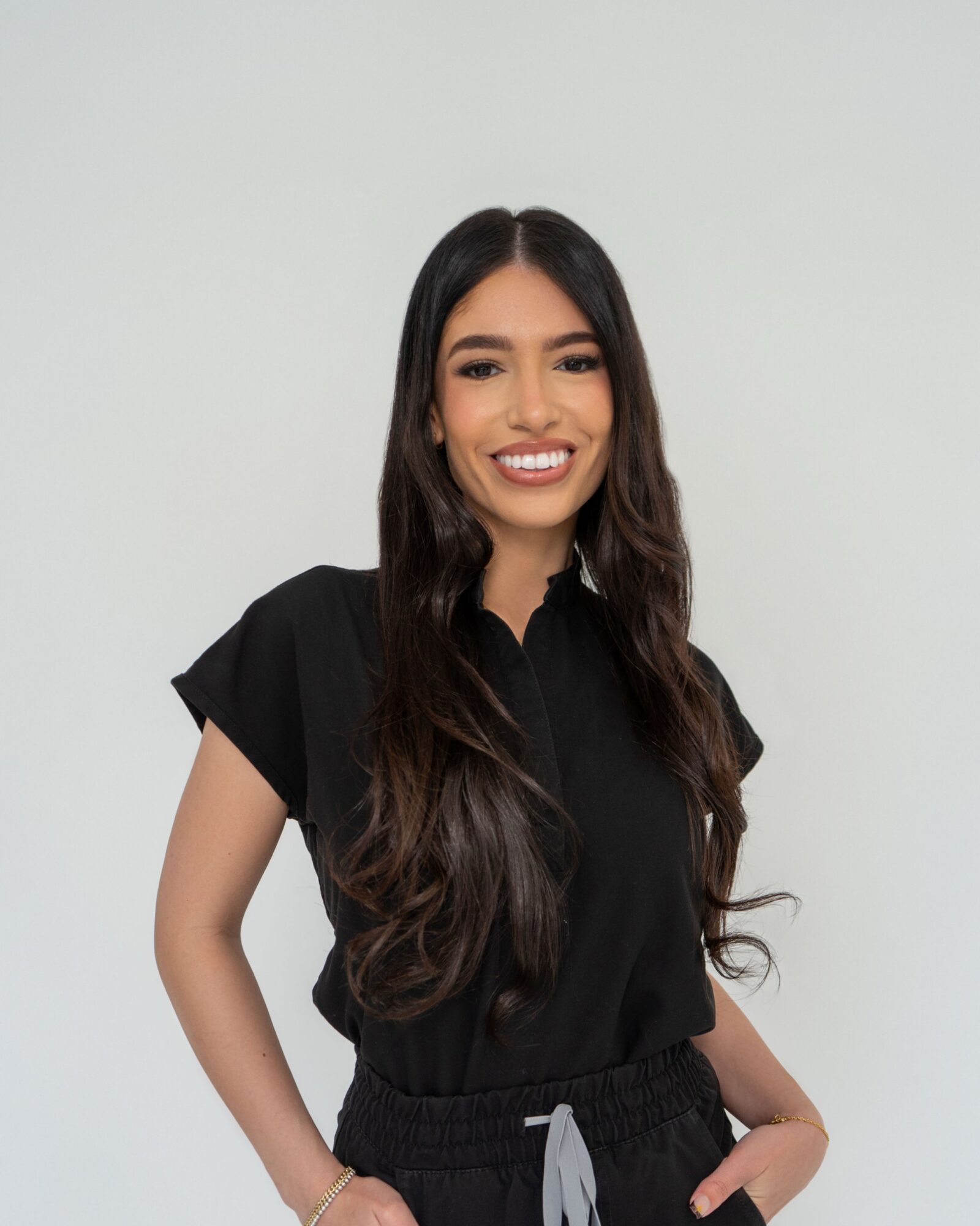 Young woman with long dark hair smiling, wearing a black shirt, standing against a plain light background.