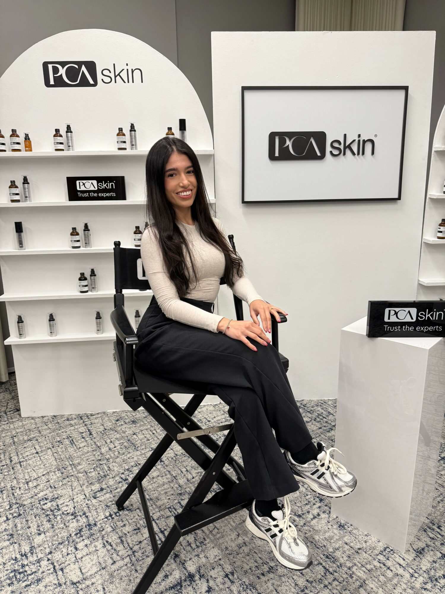 Woman sitting on a black chair in a skincare store, holding a phone, with shelves of skincare products behind her.