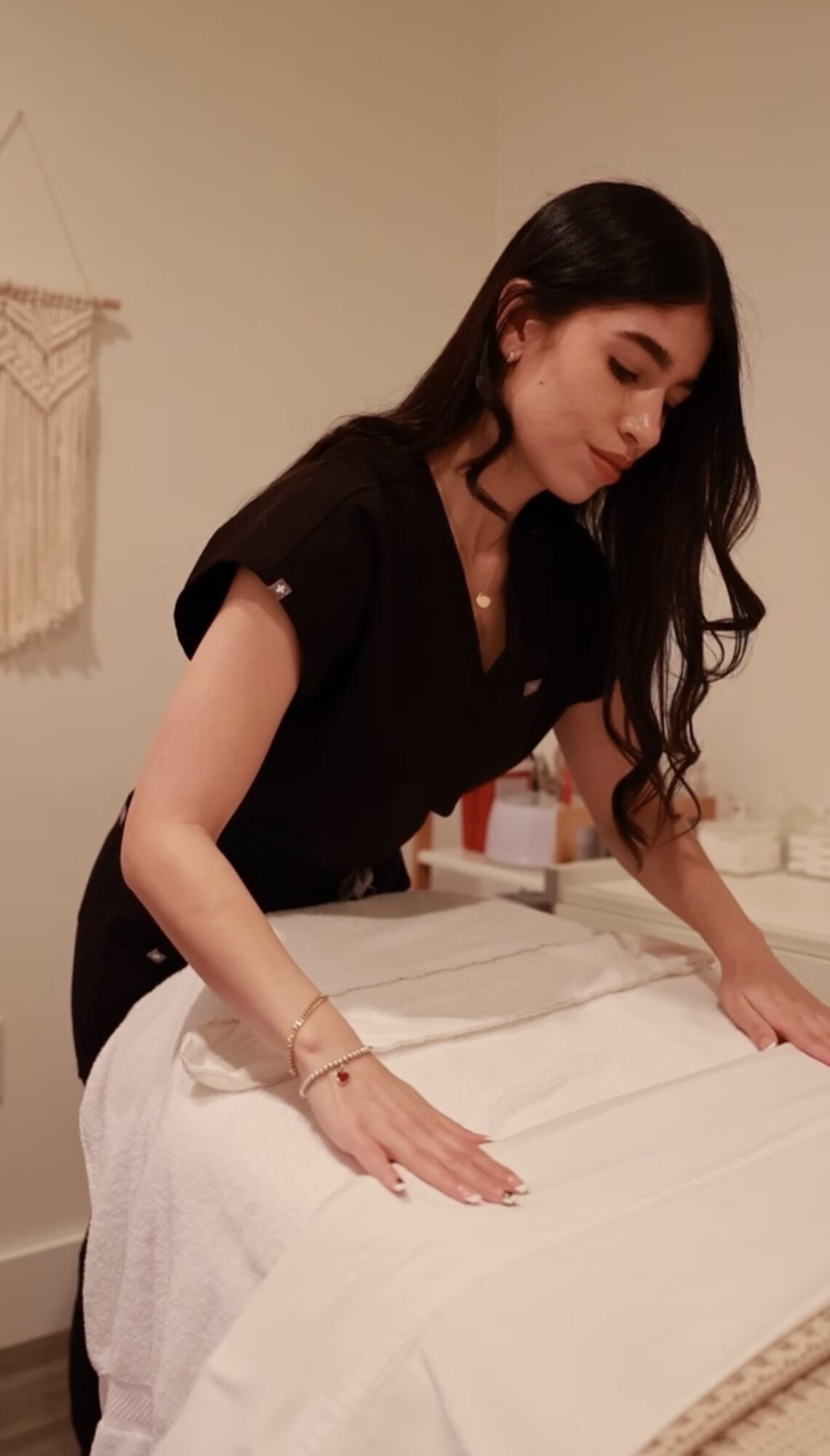 Woman with long dark hair leaning over a massage table, preparing for a treatment in a softly lit room.