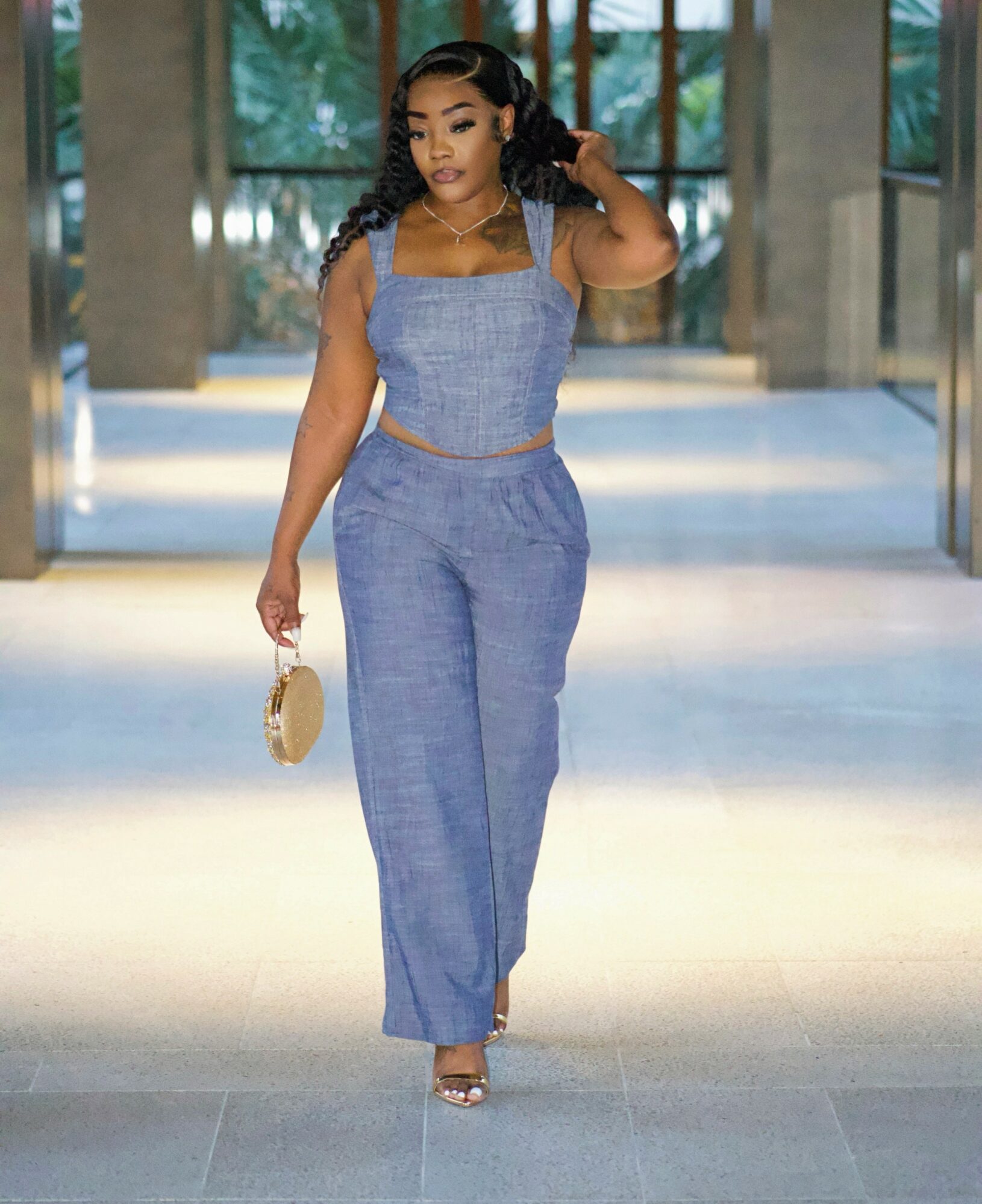 Woman in blue outfit walking indoors, holding a small bag, with large windows and trees outside in background.