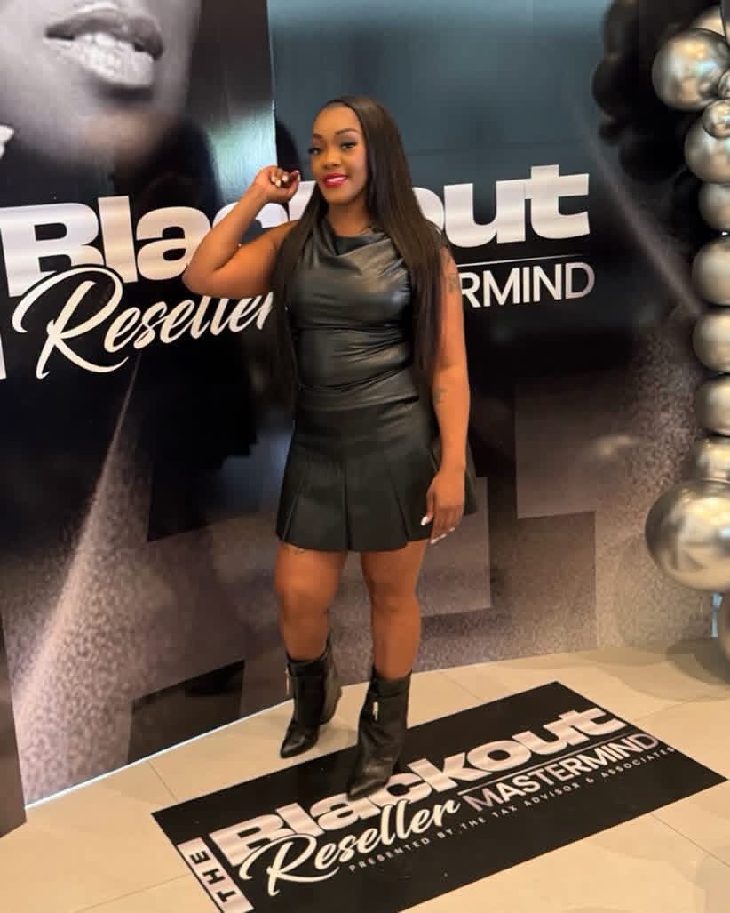 Woman in black dress and boots standing in front of a promotional backdrop, smiling and holding a small object.