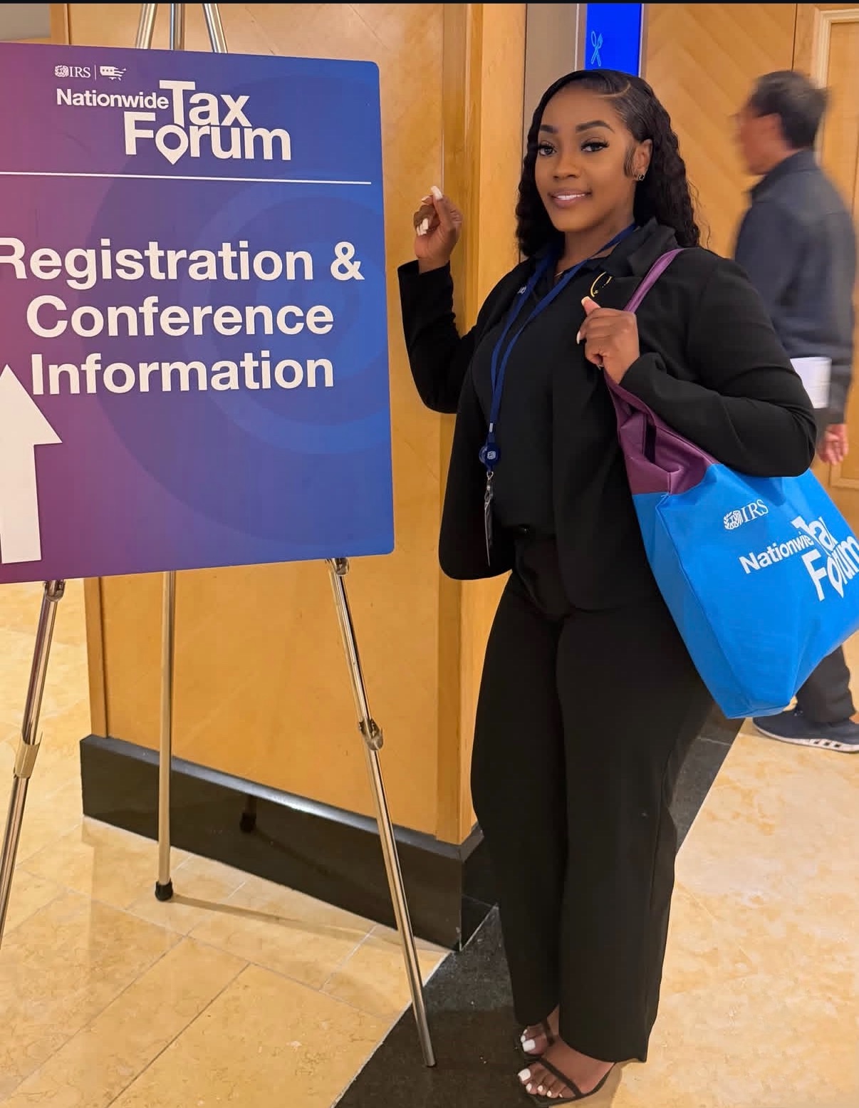 Young woman with a backpack and tote bag standing next to a conference sign, smiling, in an indoor setting.