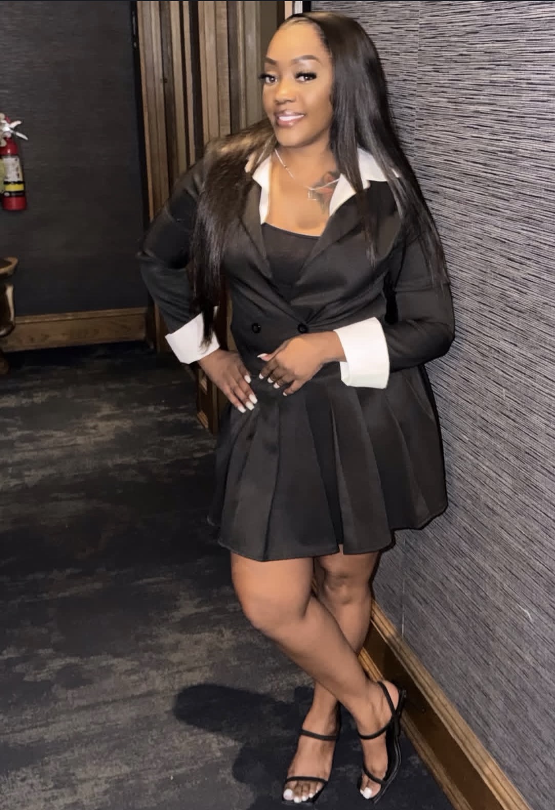 Woman in black dress with white cuffs, smiling, standing indoors against textured wall, wearing high heels.