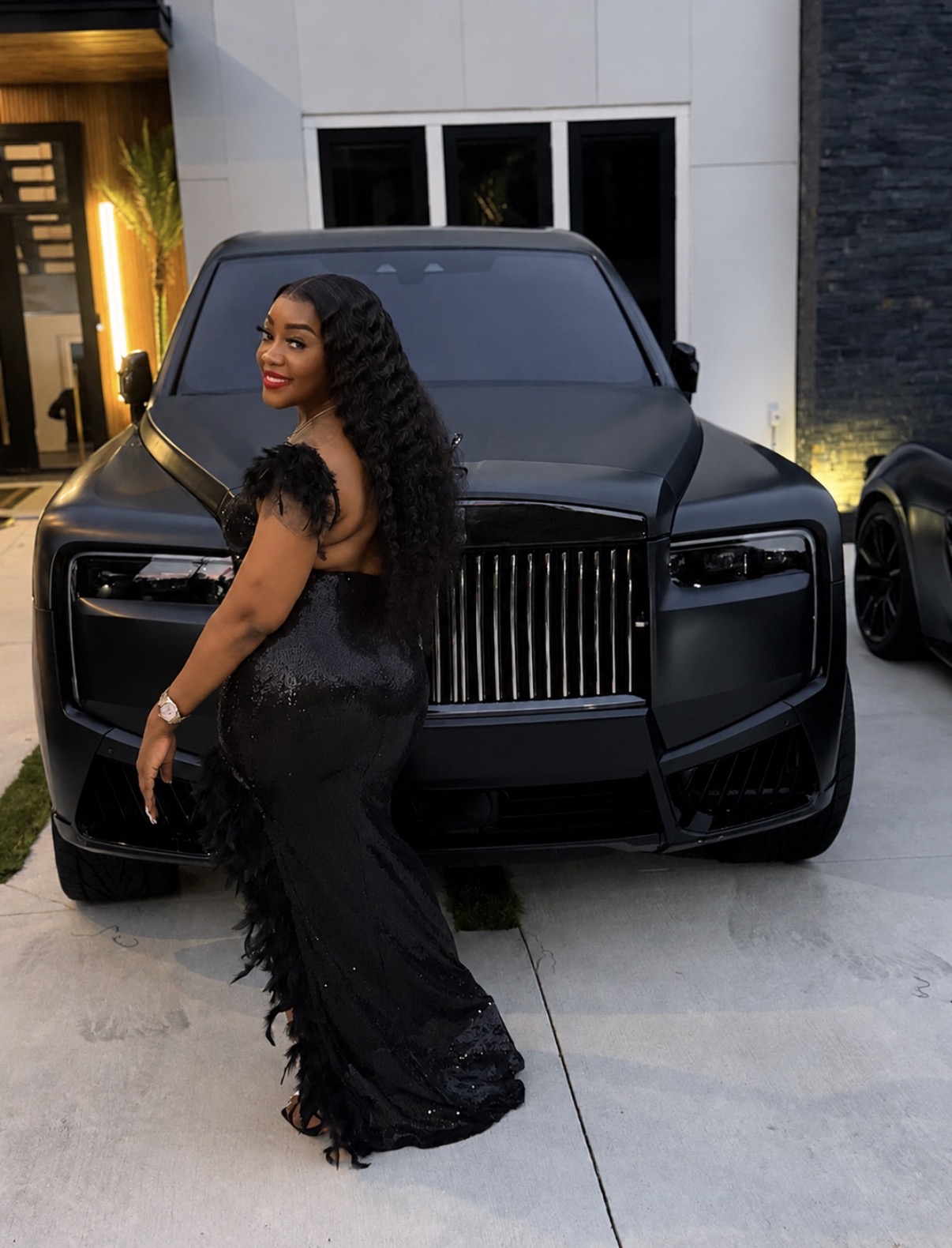 Woman in a black dress with curly hair standing in front of a black luxury car outside a modern building.