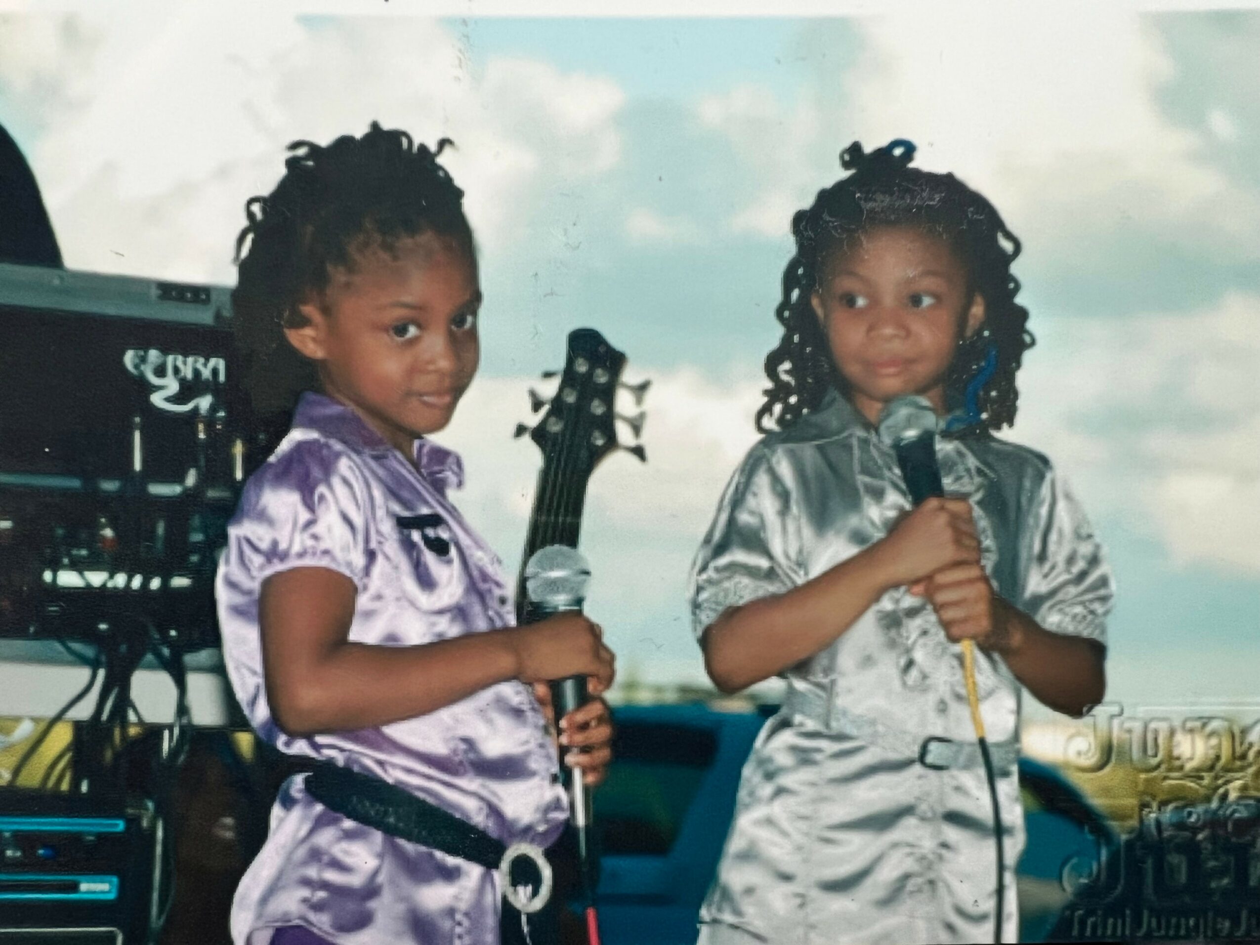 Two young girls with curly hair holding microphones, standing outdoors with equipment in the background.