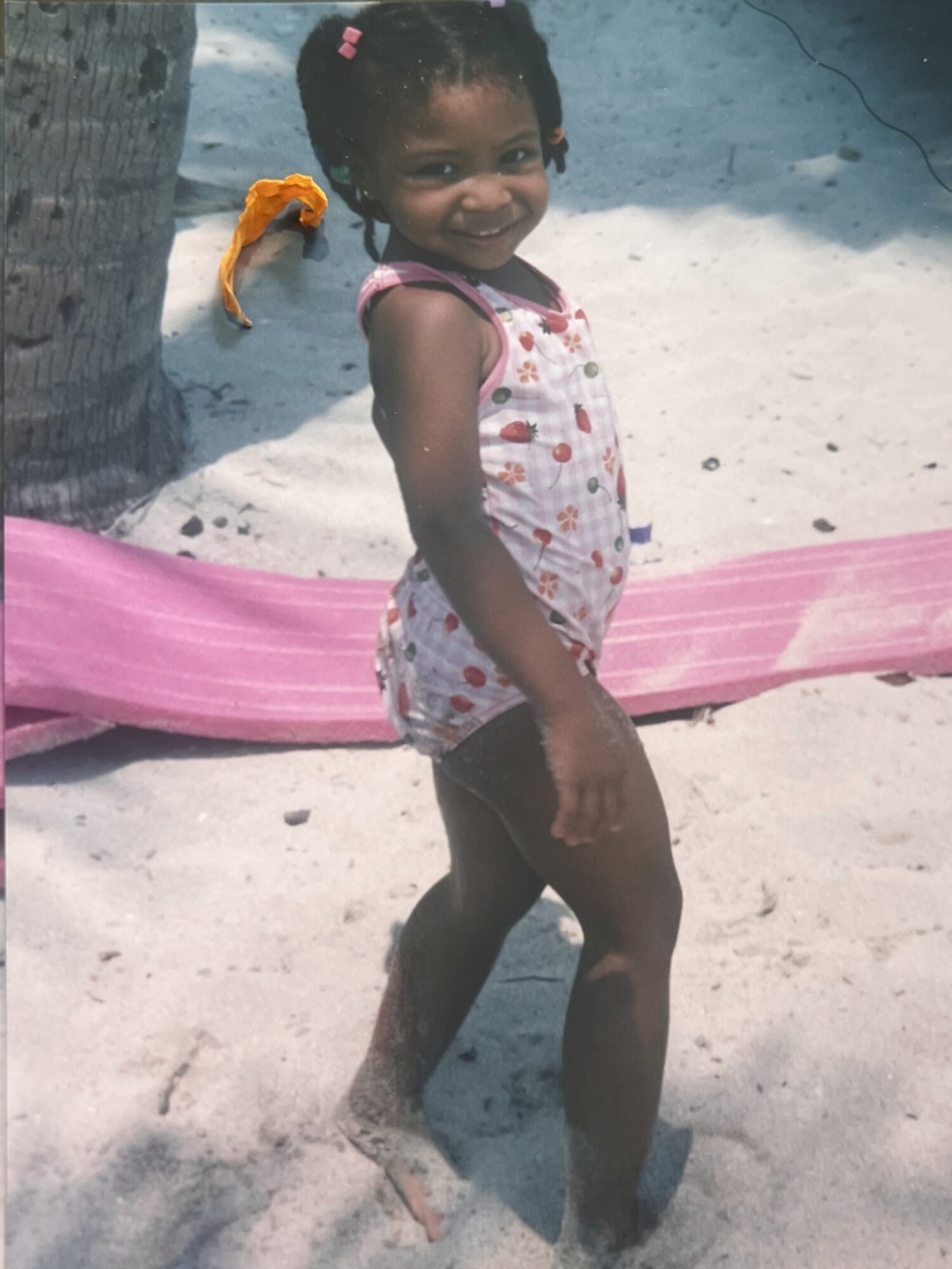 Young girl with curly hair and a pink hair clip, smiling, standing on sand near a pink hammock and a tree.