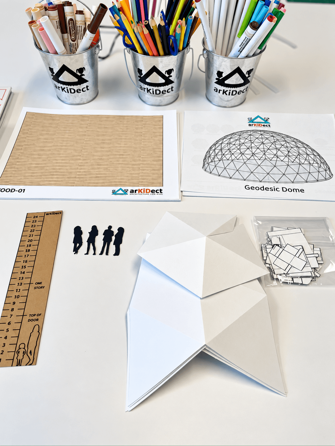 Colored pencils, pens, and markers in cups, a cardboard sheet, geodesic dome diagram, paper model, and ruler on a white surface.