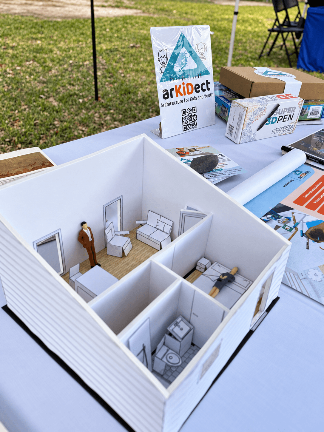 Miniature room model with furniture and a person figure inside, displayed on a table outdoors, with informational signs nearby.