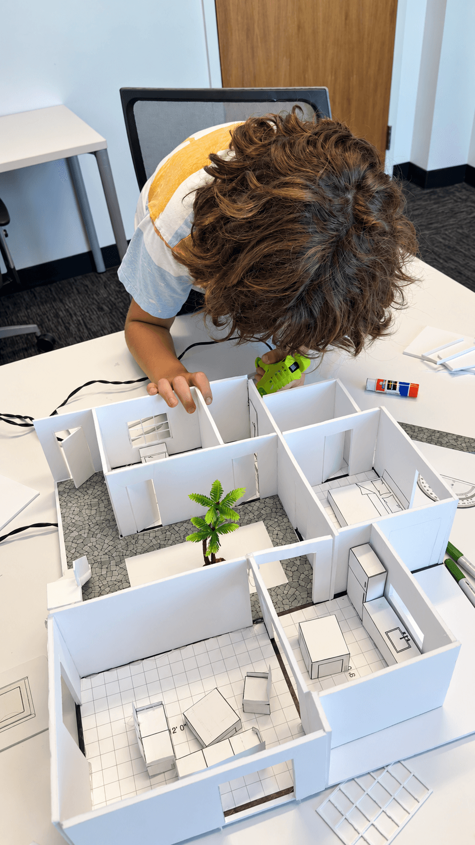 Child with curly hair leaning over a table with white model rooms and small green plants, working on a project.