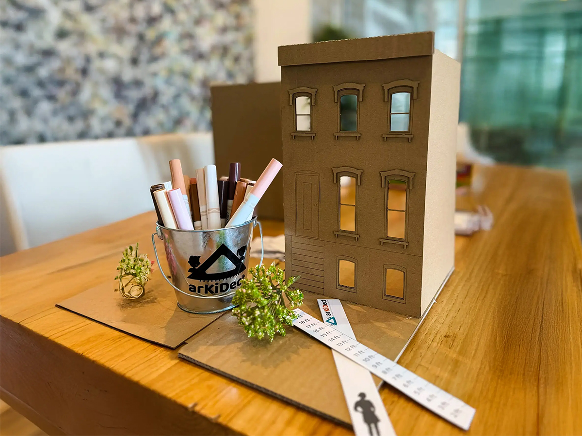 Cardboard model of a building, a container with pens, a measuring tape, and small plants on a wooden table.