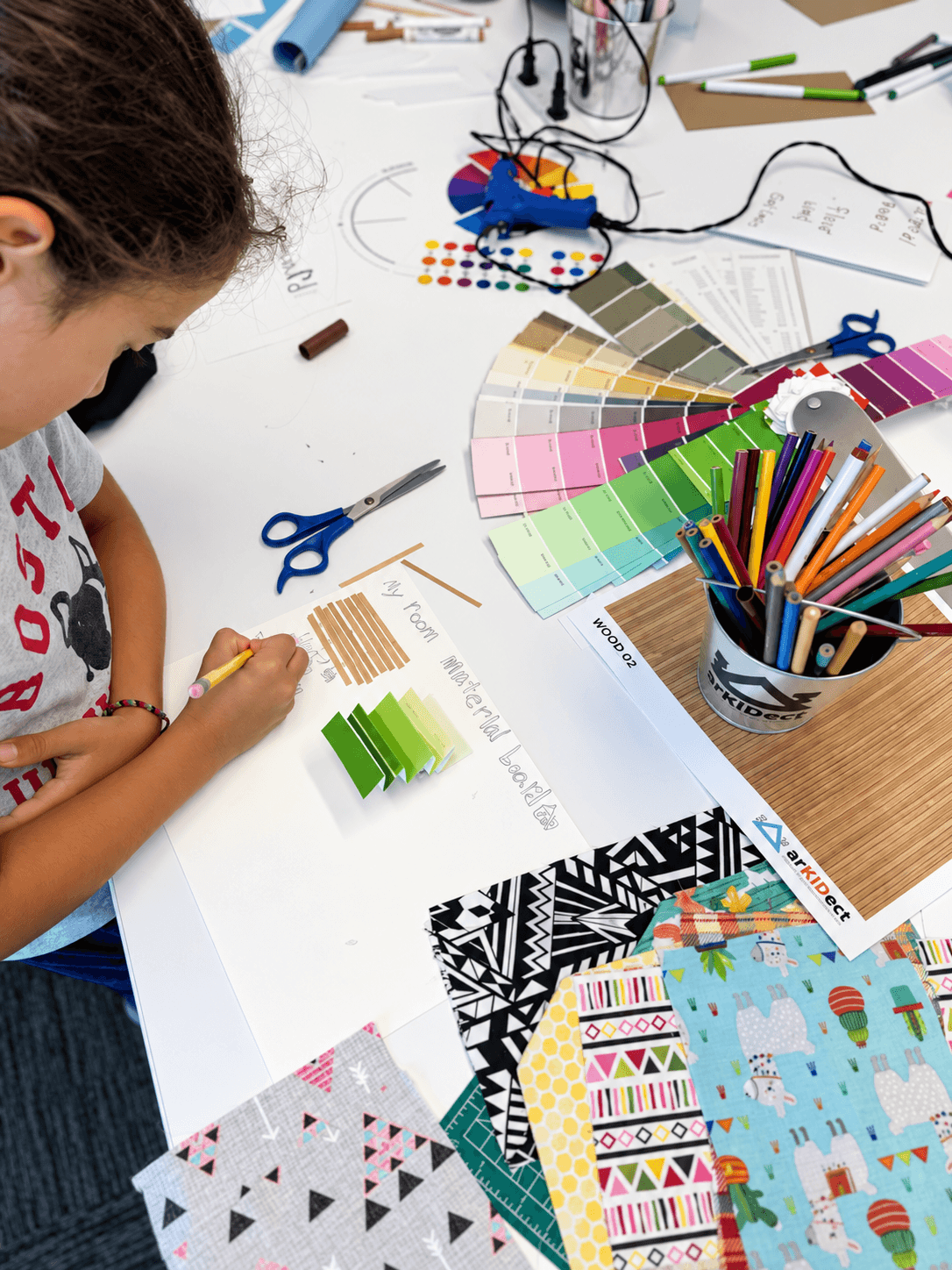 Child working on a craft project with color swatches, fabric samples, and art supplies on a white table.