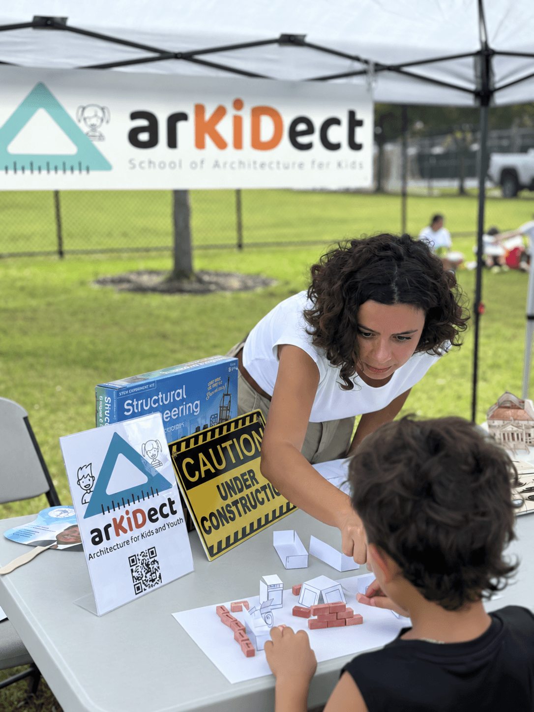 Woman and child at table with construction signs and models outdoors, under a canopy, in a park setting.