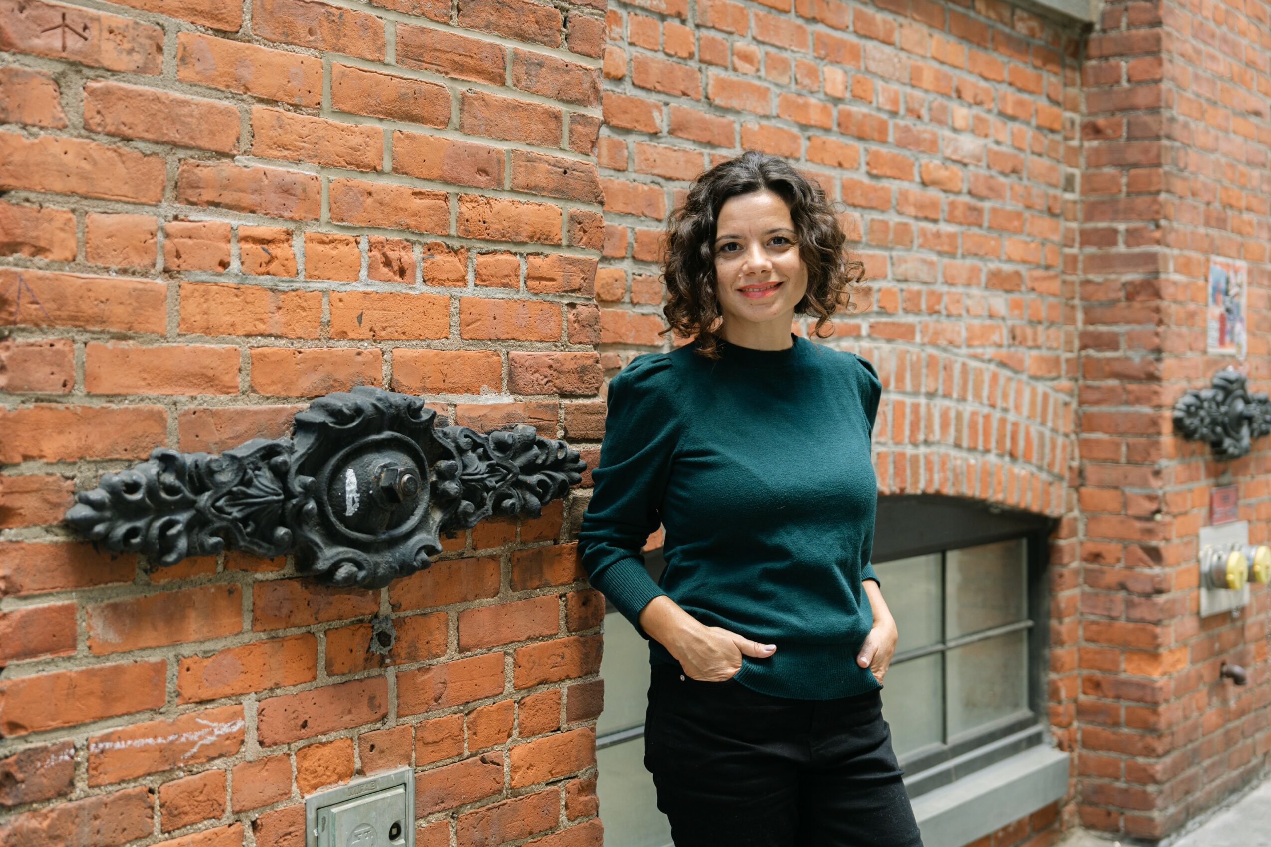 Woman with curly hair standing against a brick wall, smiling, wearing a dark green top and black pants.