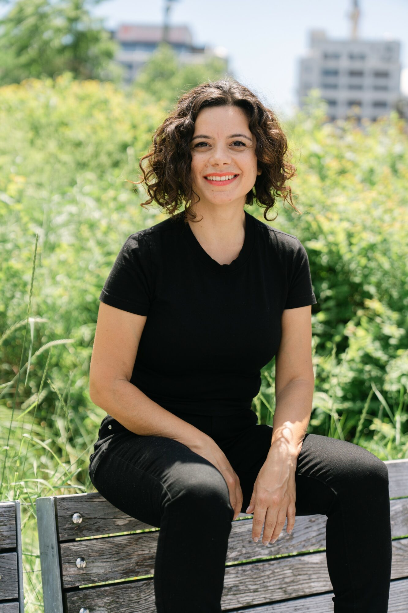 Woman with curly hair smiling outdoors, sitting on a wooden bench, wearing a black shirt and pants, greenery in background.