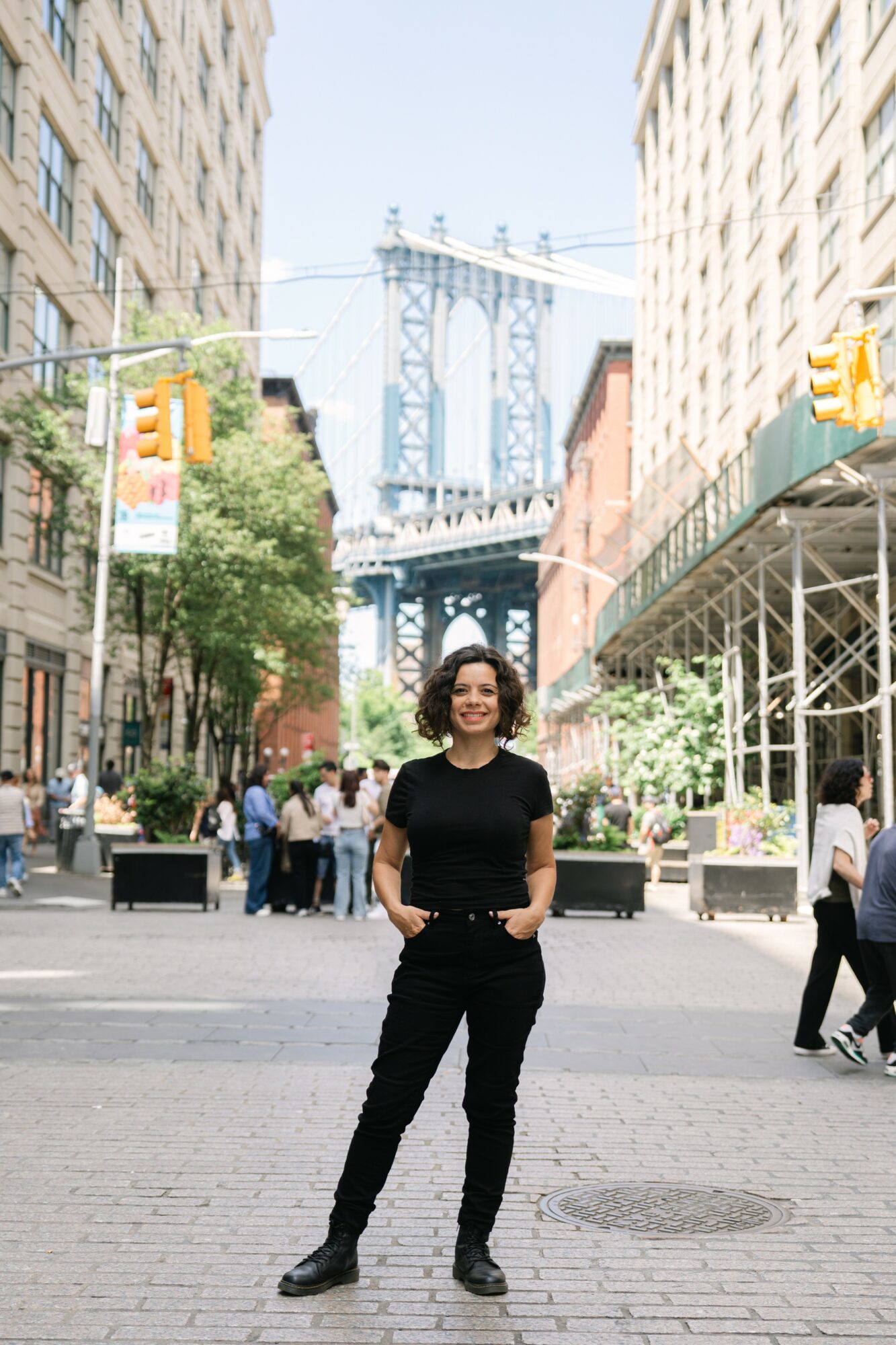 Woman standing in city street with bridge in background, smiling, wearing black outfit, surrounded by pedestrians and buildings.
