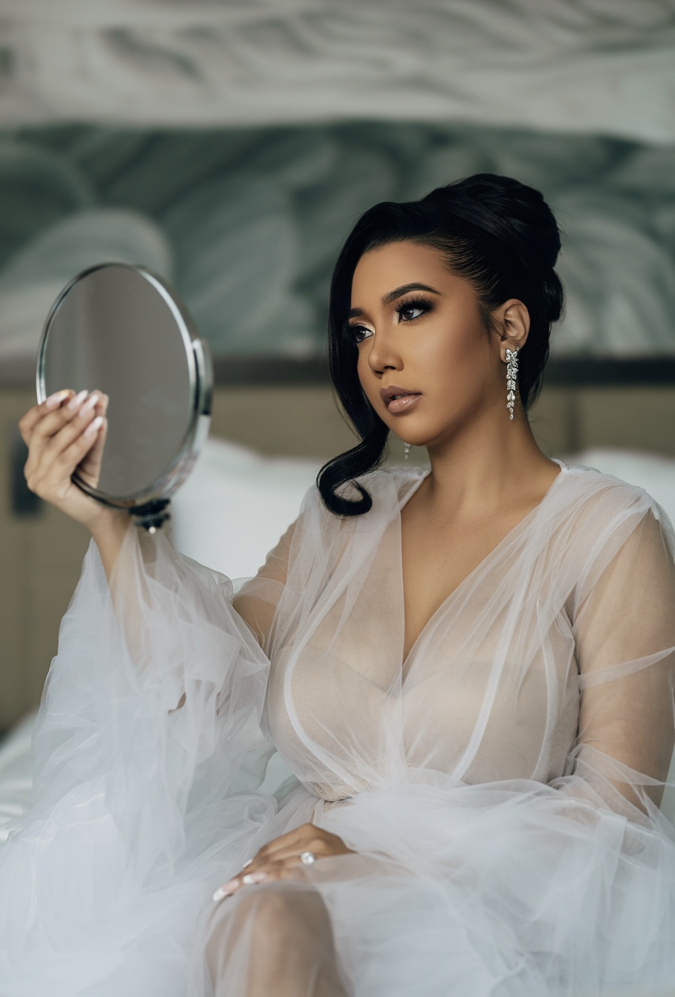 Woman with dark hair in an elegant dress holding a mirror, looking to the side, with a blurred background.