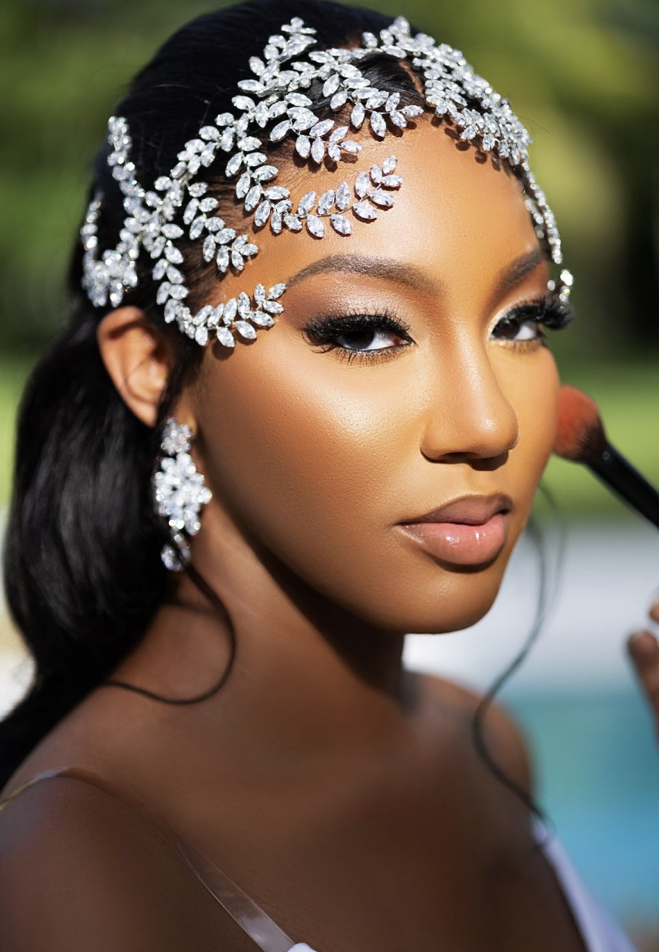 Close-up of a woman with elaborate pearl and leaf headpiece, earrings, and makeup, outdoors with blurred background.