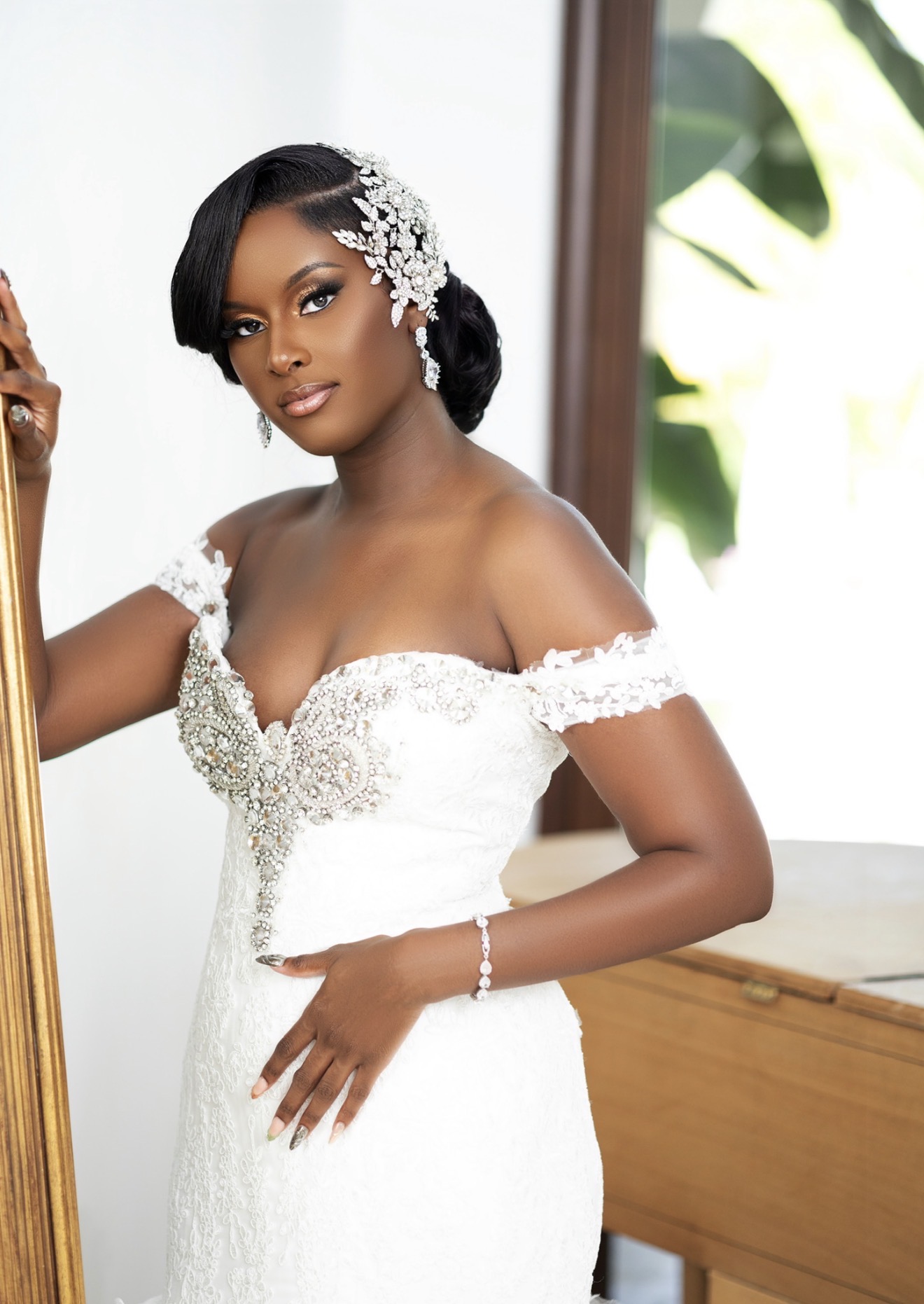 Woman in wedding dress with floral headpiece, standing indoors near a window with greenery outside.