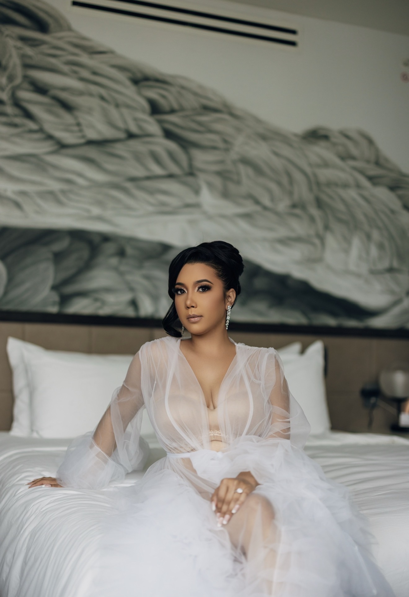 Woman in a sheer white robe sitting on a bed with white pillows, large abstract artwork behind her, neutral room decor.
