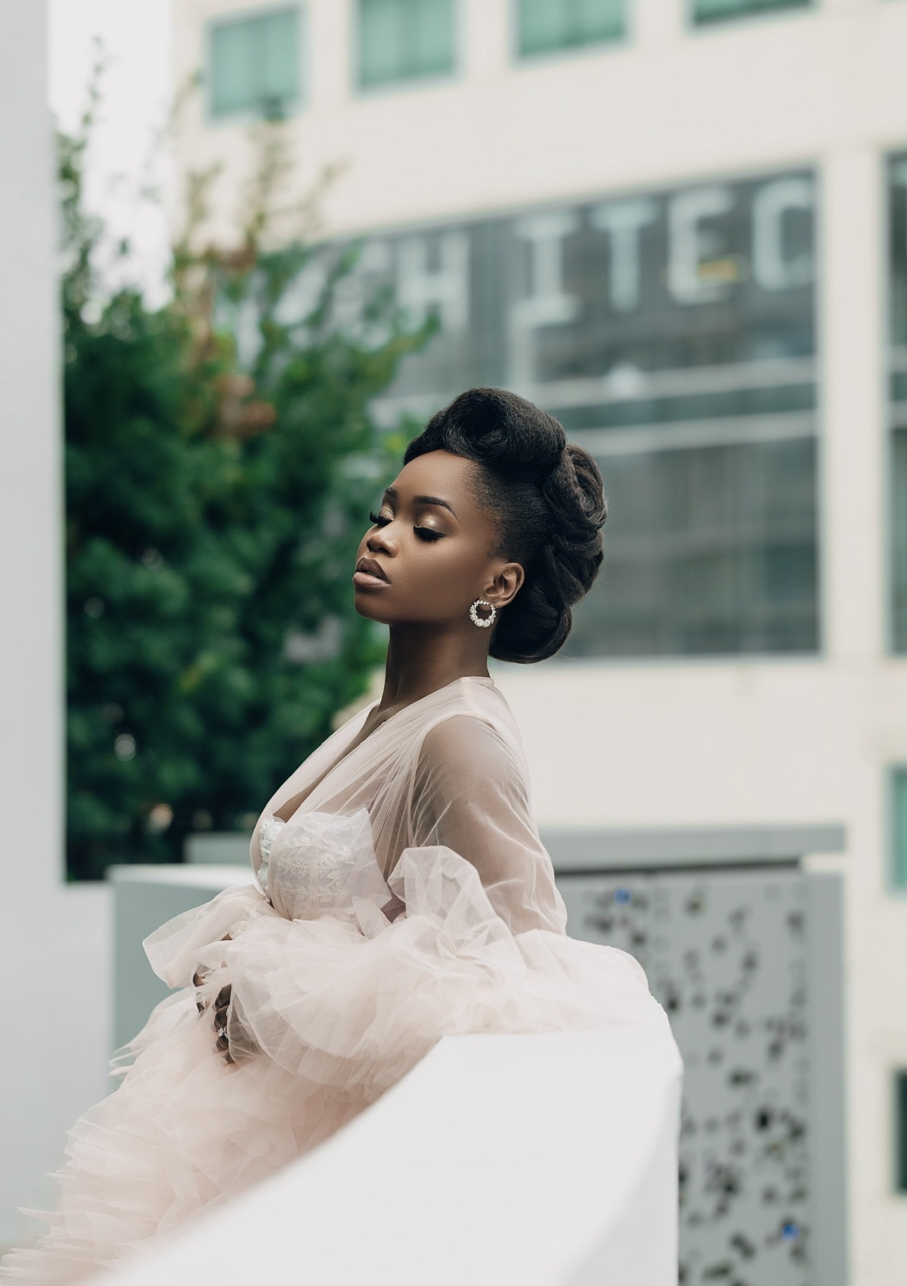 A woman in a light-colored dress with sheer sleeves stands outdoors with a modern building and greenery in the background.