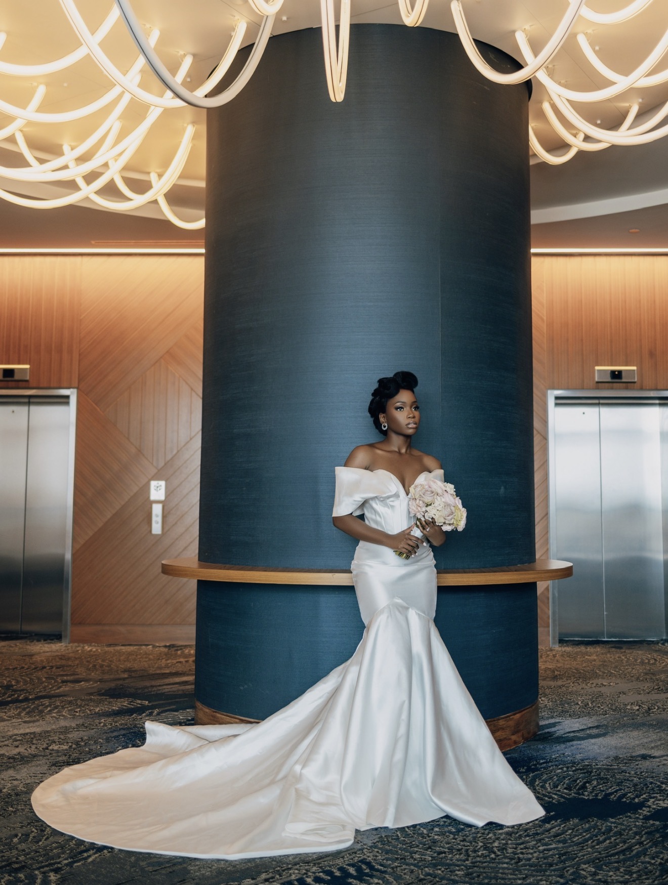 Woman in wedding dress holding bouquet stands in front of a large blue column and modern decor.