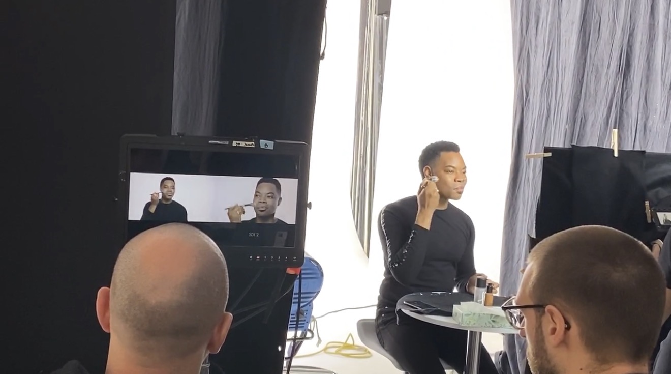 Person speaking into a microphone during a recording session, with two people in the foreground and a camera recording the scene.