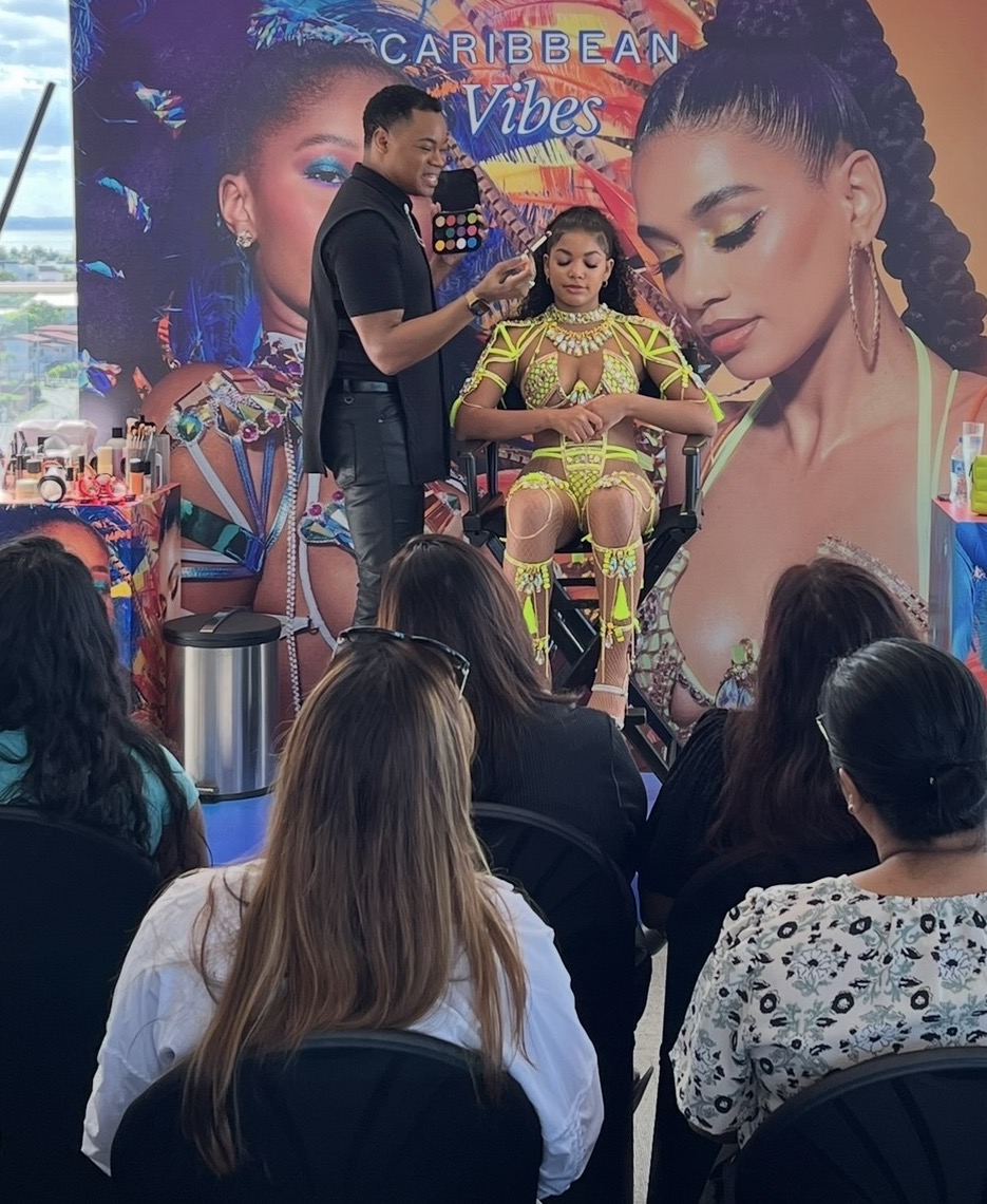 A woman receives a beauty treatment on stage with a large Caribbean-themed backdrop, audience watching attentively.