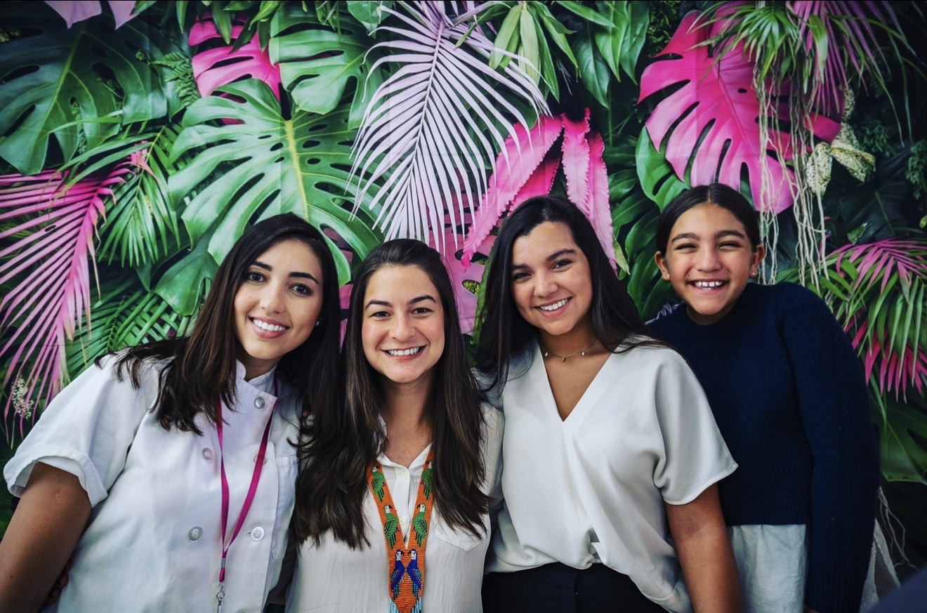Four smiling women stand in front of a vibrant tropical leaf background.