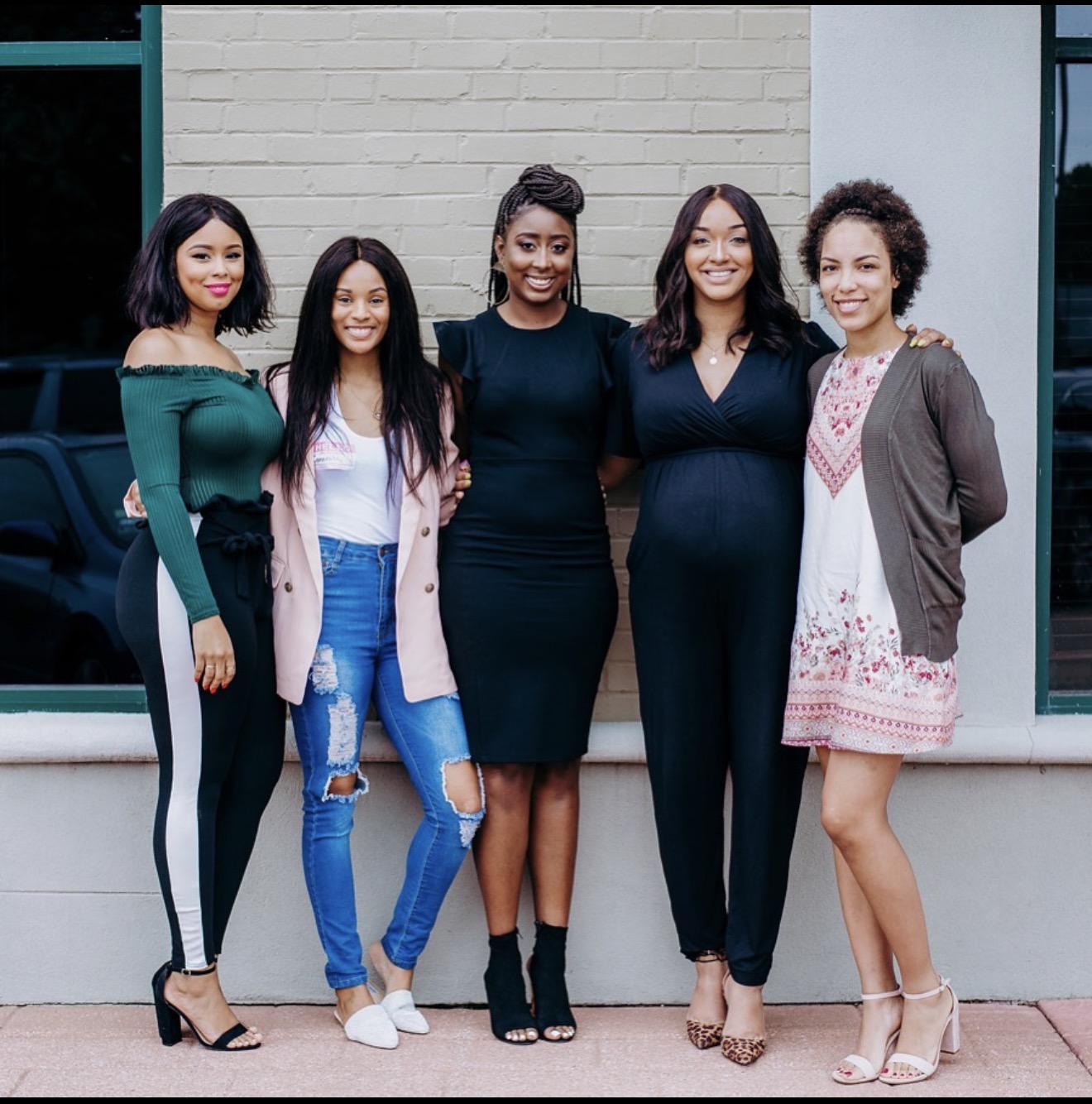 Five women standing outdoors in front of a building, smiling, with arms around each other, dressed in casual and stylish clothing.