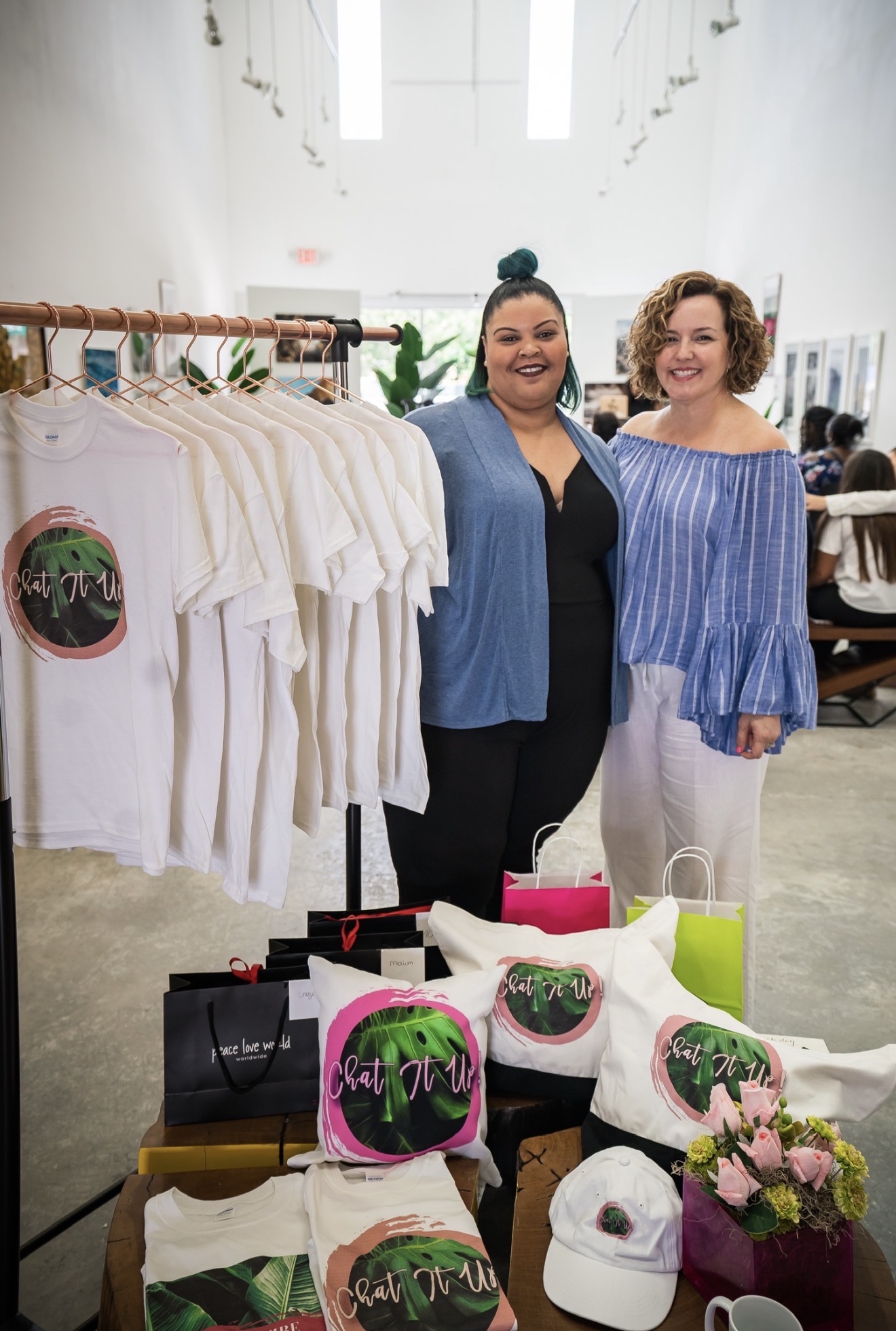 Two women stand inside a store with clothing racks and tables displaying hats and accessories, smiling at the camera.