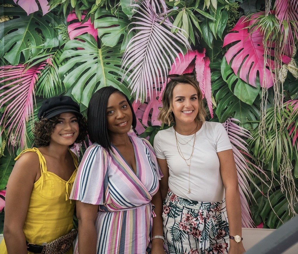 Three women standing in front of a vibrant pink and green tropical leaf backdrop, smiling and posing for the photo.