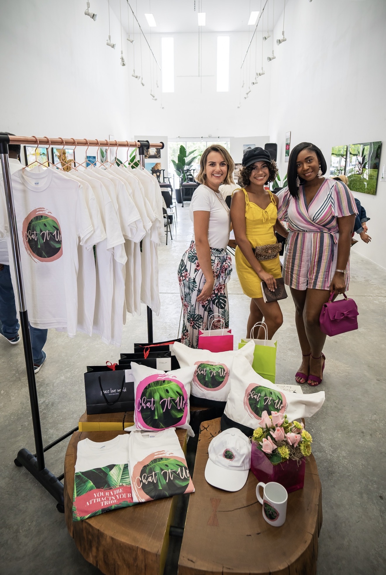 Three women stand together inside a store with white walls and clothing racks, smiling at the camera. A table with hats, bags, and flowers is in front.