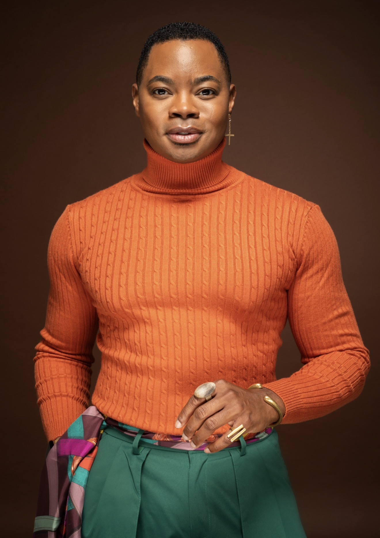 Woman with short hair wearing an orange turtleneck sweater and green pants, standing against a brown background.