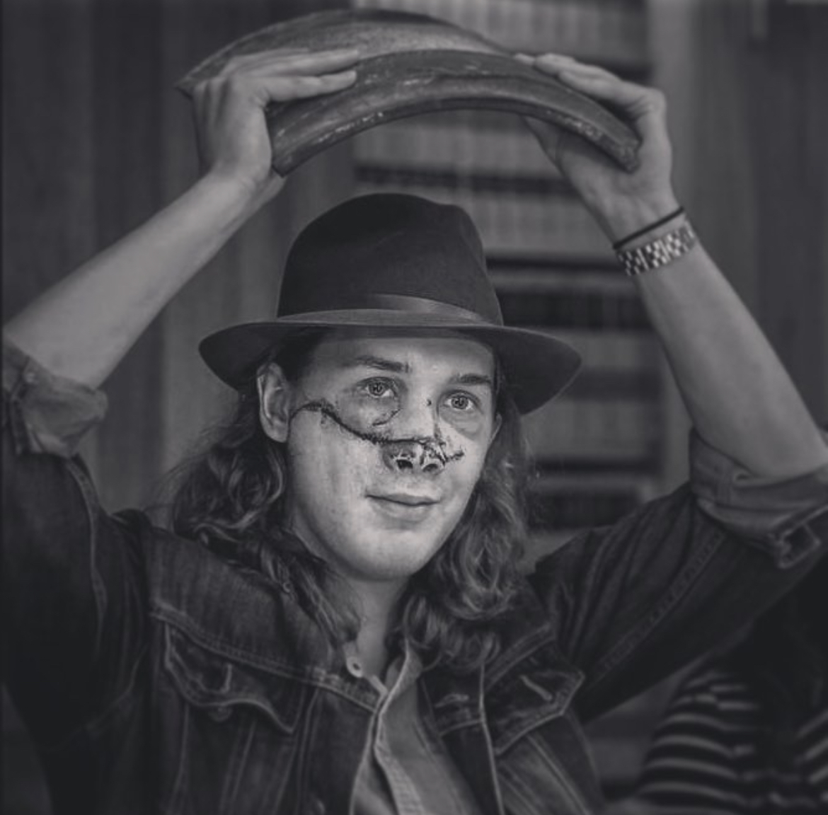 Person with long hair, wearing a hat and denim jacket, holds a large object above their head, smiling. Word count: 20
