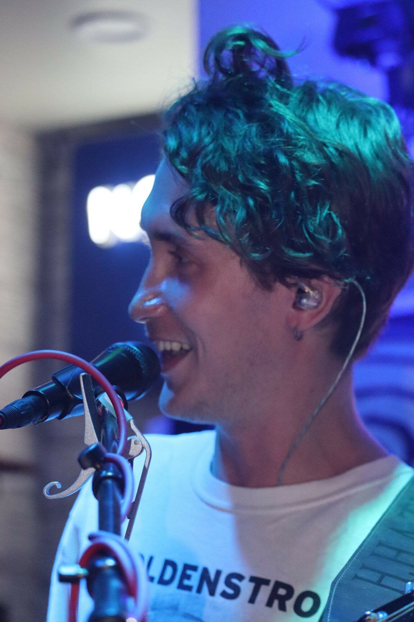 Person with curly hair smiling, wearing earphones, speaking into a microphone, in a blue-lit environment.
