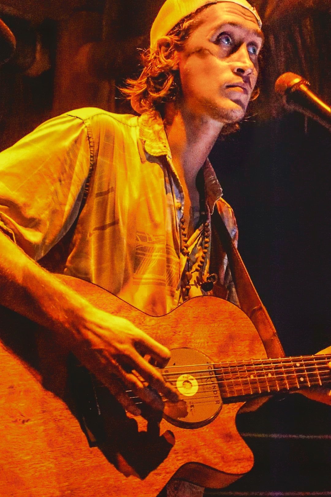Person with long hair wearing a yellow shirt, playing an acoustic guitar on stage, with a microphone nearby.