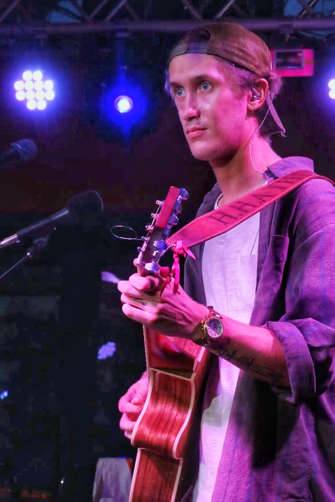 Young person with long hair, wearing a headband, playing an acoustic guitar on stage with purple and blue lights.