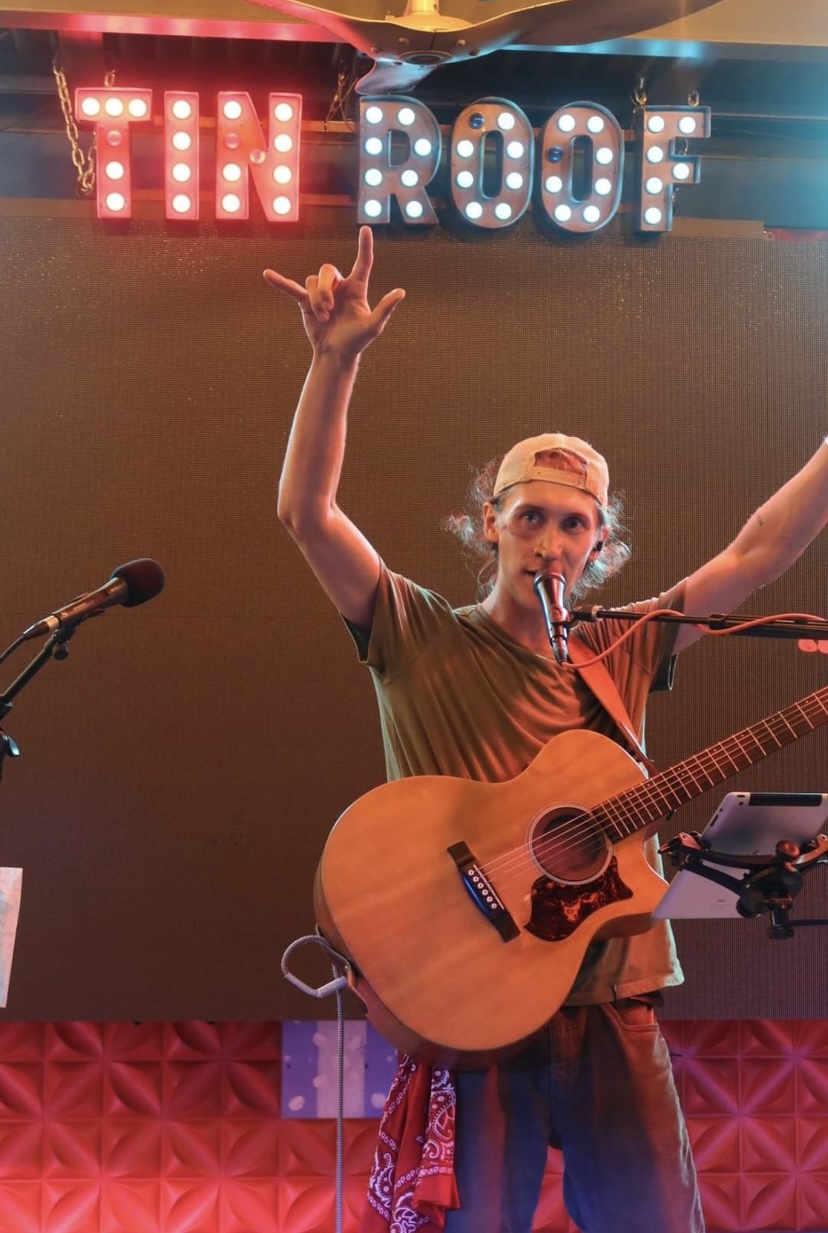 Person playing guitar on stage with a sign reading 'TIN ROOF' above, raising one hand, wearing a cap, microphone nearby.