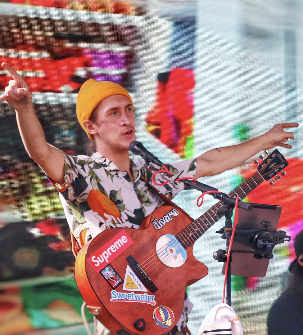 Person wearing a yellow hat and floral shirt, singing into a microphone, playing guitar with stickers, in a store setting.