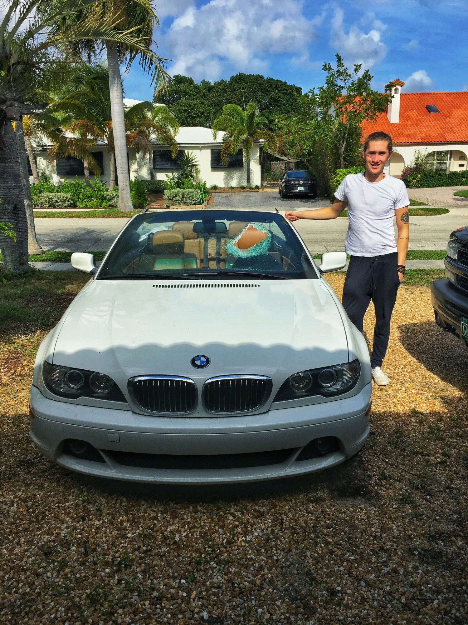 Person standing next to a white BMW convertible car in a parking lot with trees and houses in the background.