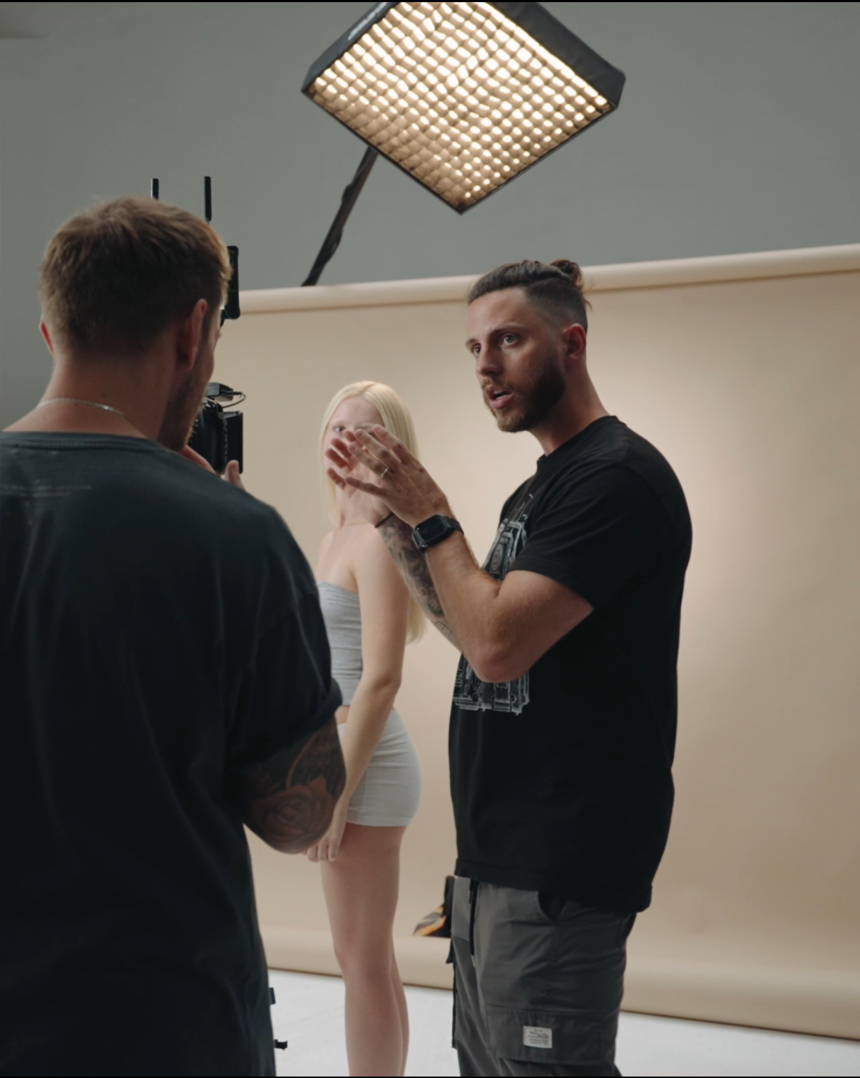 Two men in conversation in a photography studio with a woman in the background, soft lighting, and a beige backdrop.
